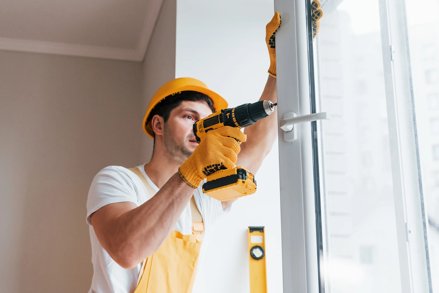 Handyman in yellow uniform installs new window by using automatic screwdriver. House renovation conception<br />